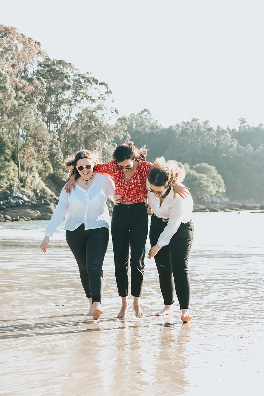 Three young women walking barefoot on the beach, enjoying a sunny day. They are dressed in casual outfits, with two wearing white shirts and one in a red polka dot top. The background features lush greenery and calm waters, creating a serene atmosphere.