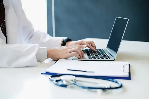 A healthcare professional in a white lab coat typing on a laptop, with a stethoscope and notepad on the table, illustrating the integration of technology in modern medicine.